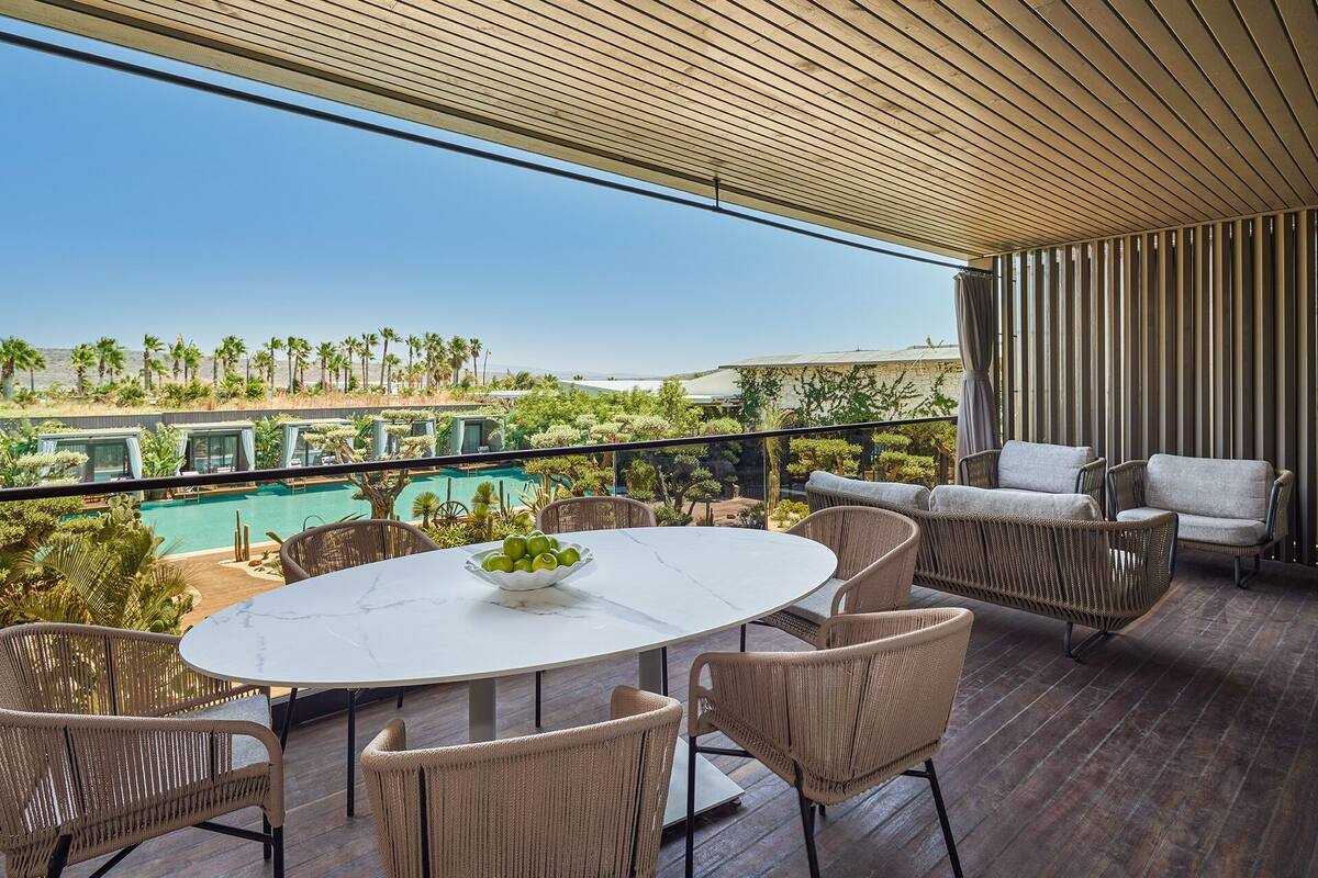 Un balcón con una mesa redonda, sillas de mimbre y vistas a una piscina y palmeras.