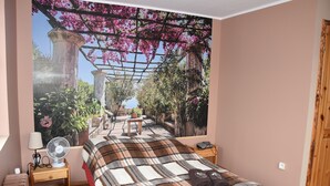 A bedroom with a large bed, a nightstand with a lamp and fan, a wooden bench, and a wall mural of a garden. - Agroturystyka u przyjaciol Soni (Zlocieniec)