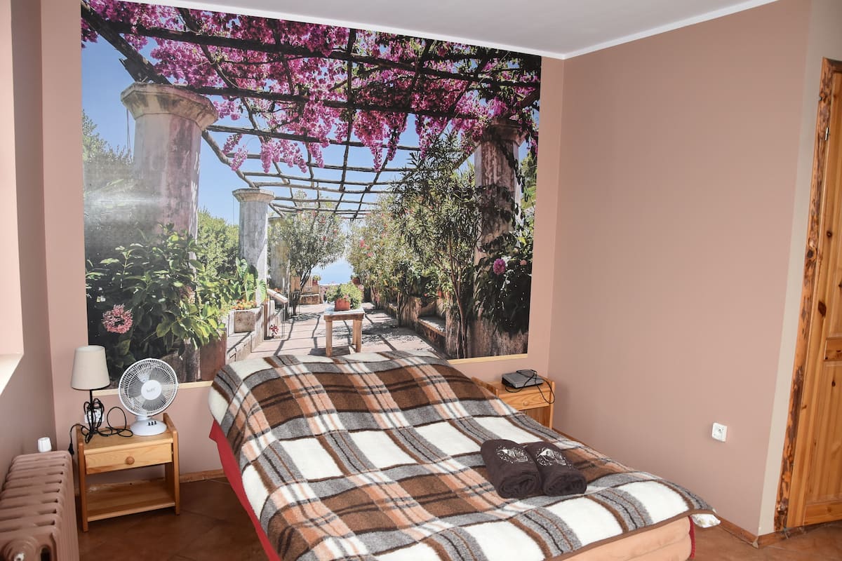 Un dormitorio con una cama grande, una mesita de noche con lámpara y ventilador, un banco de madera y un mural de jardín en la pared.