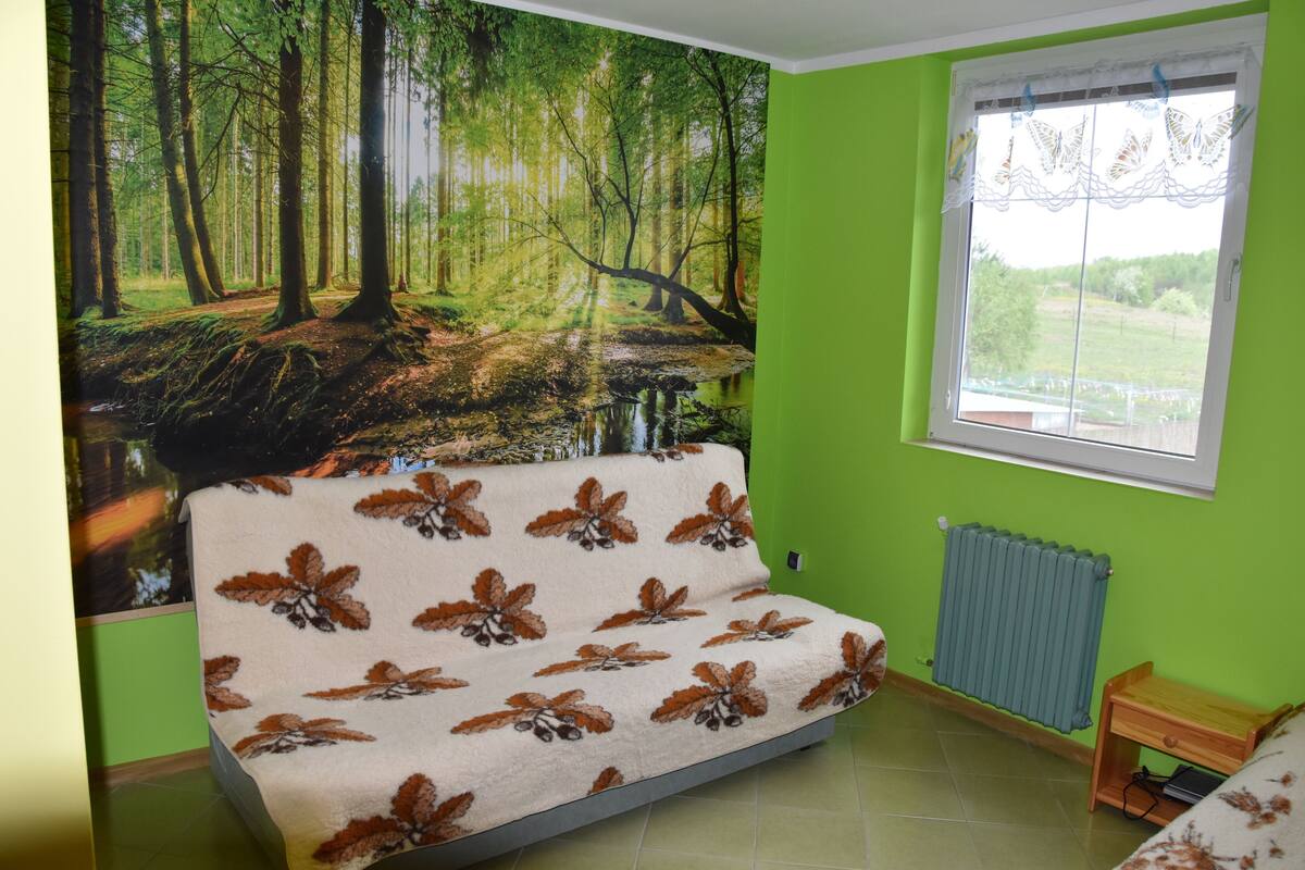 Una habitación con sofá cubierto por una manta de estampado de hojas, un radiador, una ventana con vistas a la vegetación y un mural en la pared que representa un bosque.