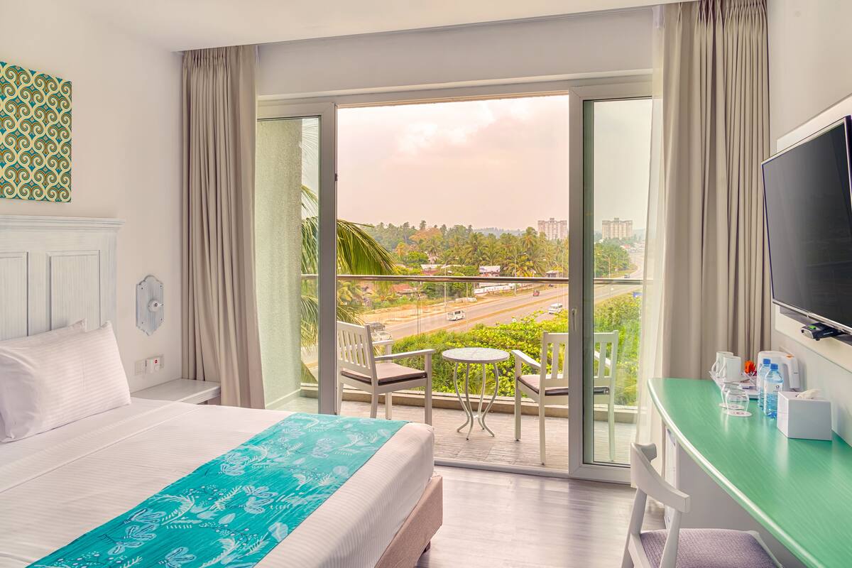 A hotel room with a bed, a desk, a TV, and a balcony with a view of the city.