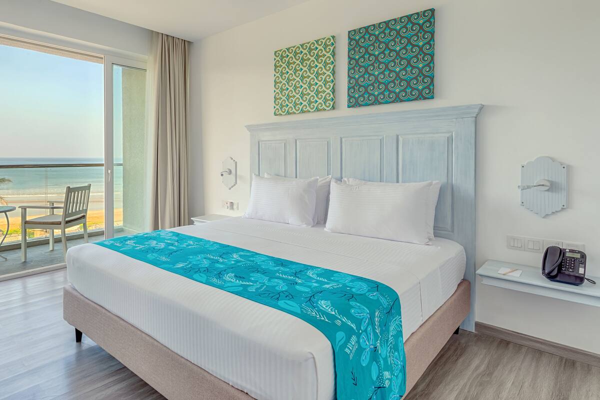 A hotel room with a large bed, a view of the beach, a balcony with a table and chairs, and a phone on a bedside table.