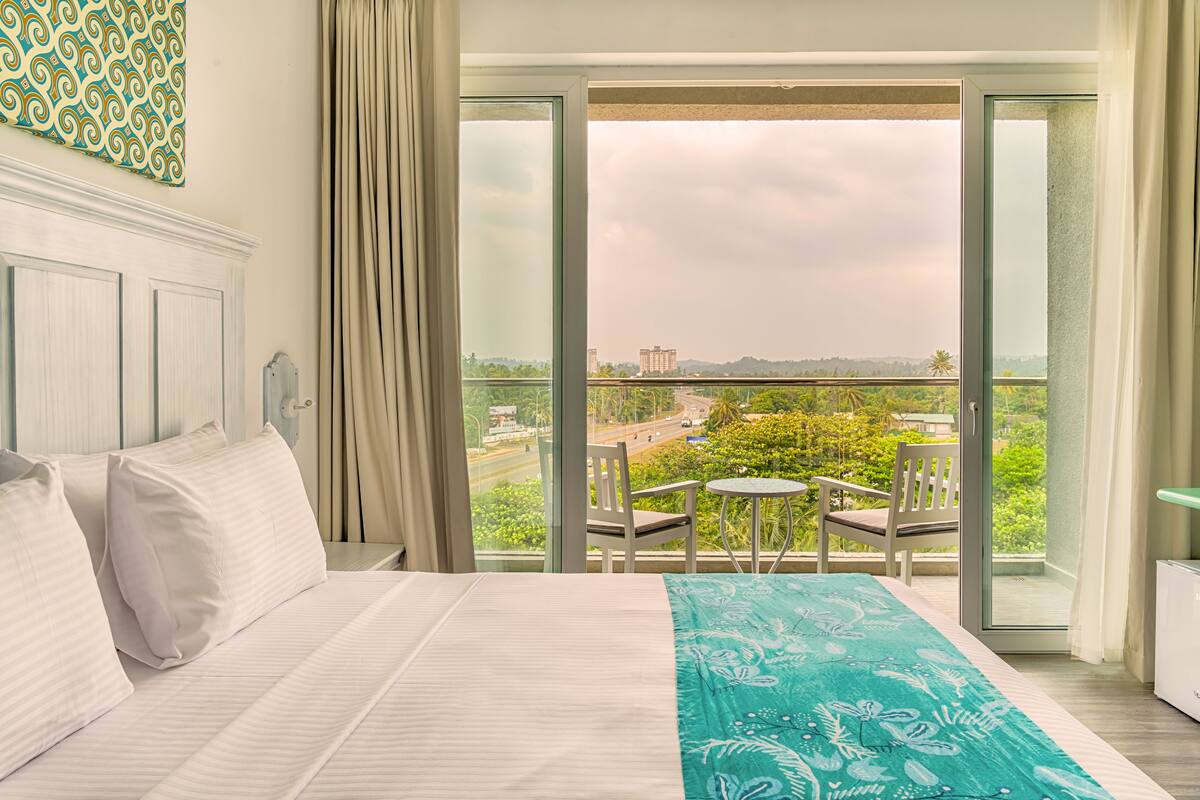 A hotel room with a large bed, a balcony with a view of the city, and a small table and chair.