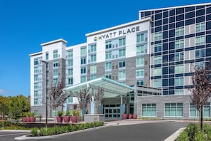 Un hotel Hyatt Place moderno con fachada de vidrio, entrada cubierta y camino de acceso arbolado.