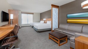 A hotel room with a wooden desk, two beds, a sofa, a TV, and a large window.