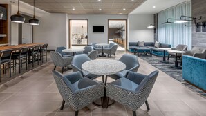 A modern lounge area with round tables and sofas, a kitchen in the background, and a large window with curtains.