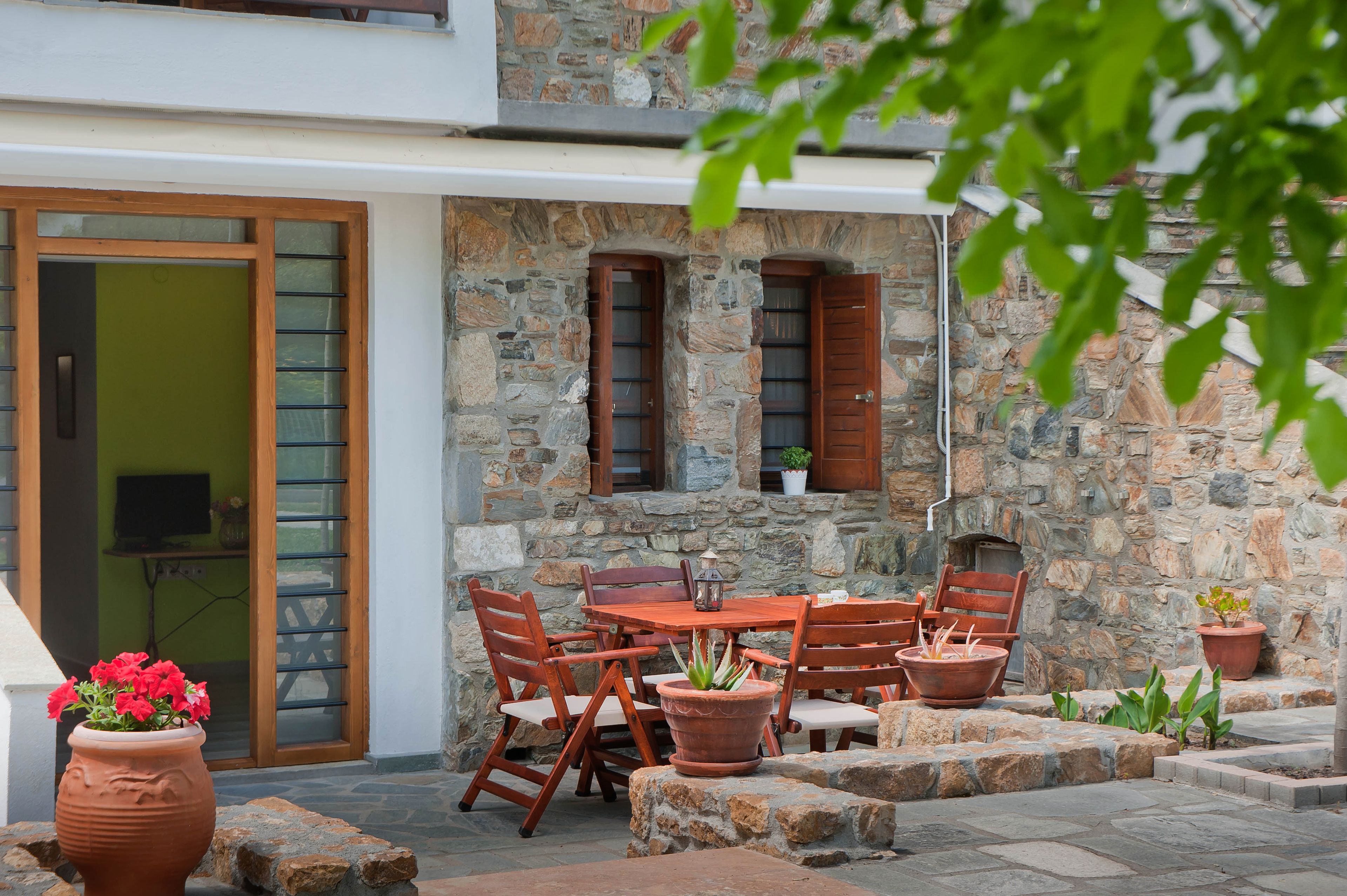 A stone building with a patio area featuring a wooden table and chairs, potted plants, and a sliding glass door.