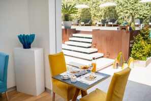 A dining area with a table set for breakfast, including plates, cups, and a bottle of orange juice. There are yellow chairs and a blue vase on a white pedestal.