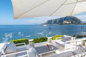 A terrace with white furniture, a table with a drink, and a view of the sea and a mountain. - Unìco Boutique Hotel d'Arte (Palermo)