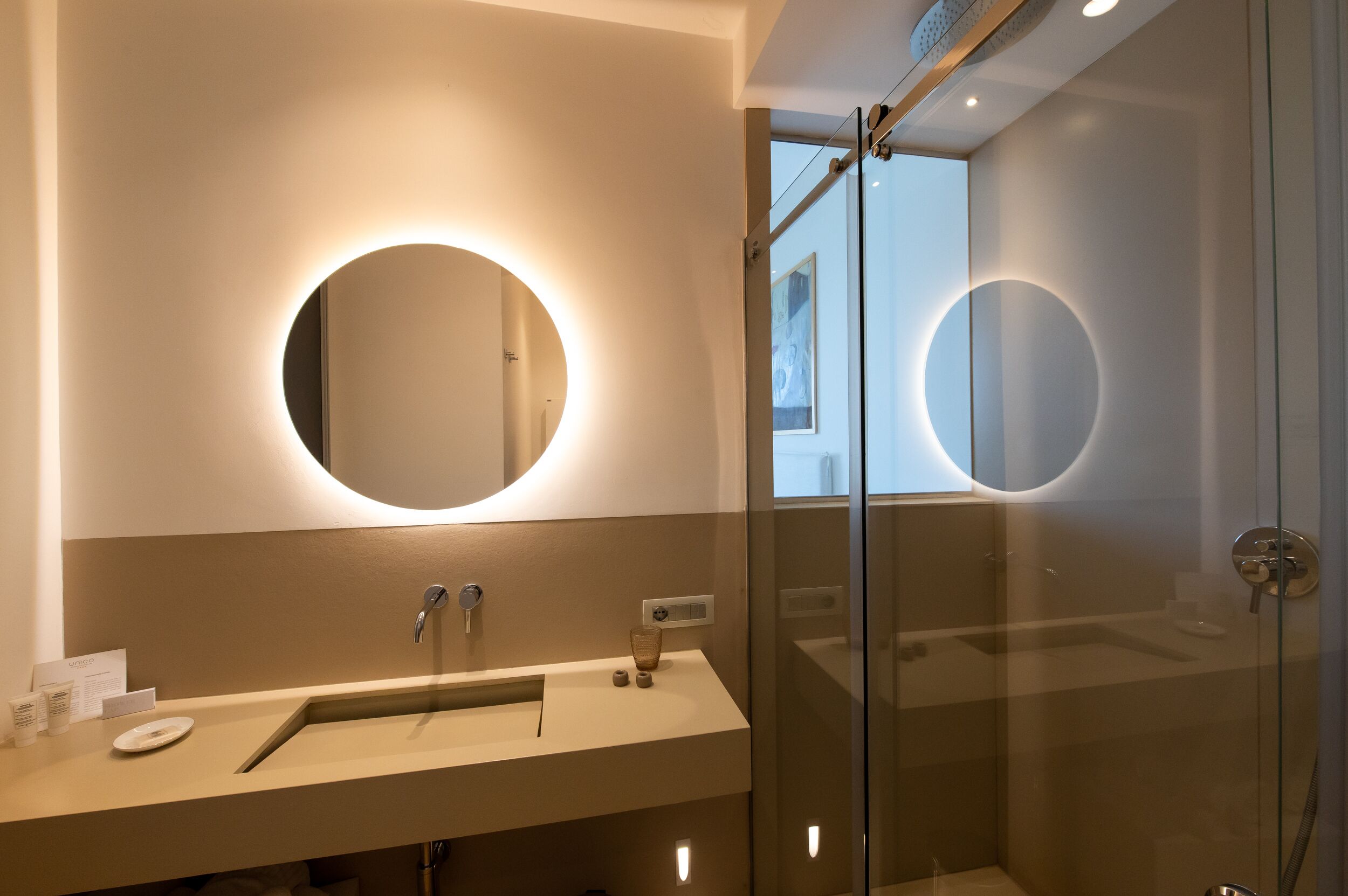 Un baño moderno con un espejo redondo, cabina de ducha de vidrio y tocador con lavabo y toallero.