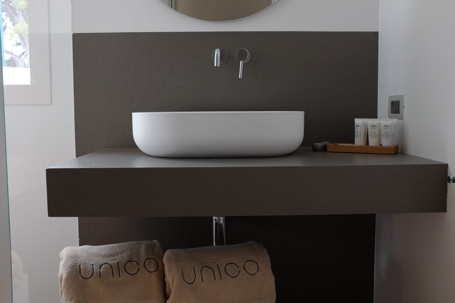 Une salle de bain moderne avec un grand lavabo sur pied, un miroir au-dessus, et deux serviettes ornées du motif « Unico ».