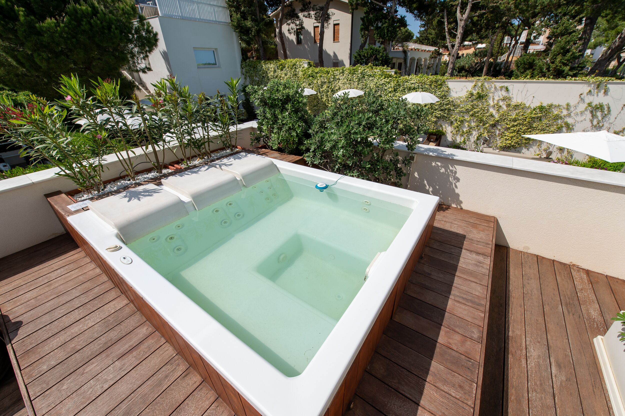 A rectangular hot tub on a wooden deck with a white border, surrounded by greenery and a white wall.