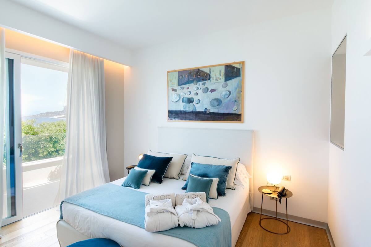A bedroom with a bed, a painting, a nightstand, and a view through a window.