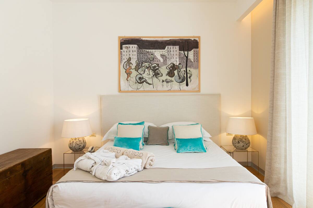 A hotel room with a bed, two bedside lamps, a painting on the wall, and a wooden bench.