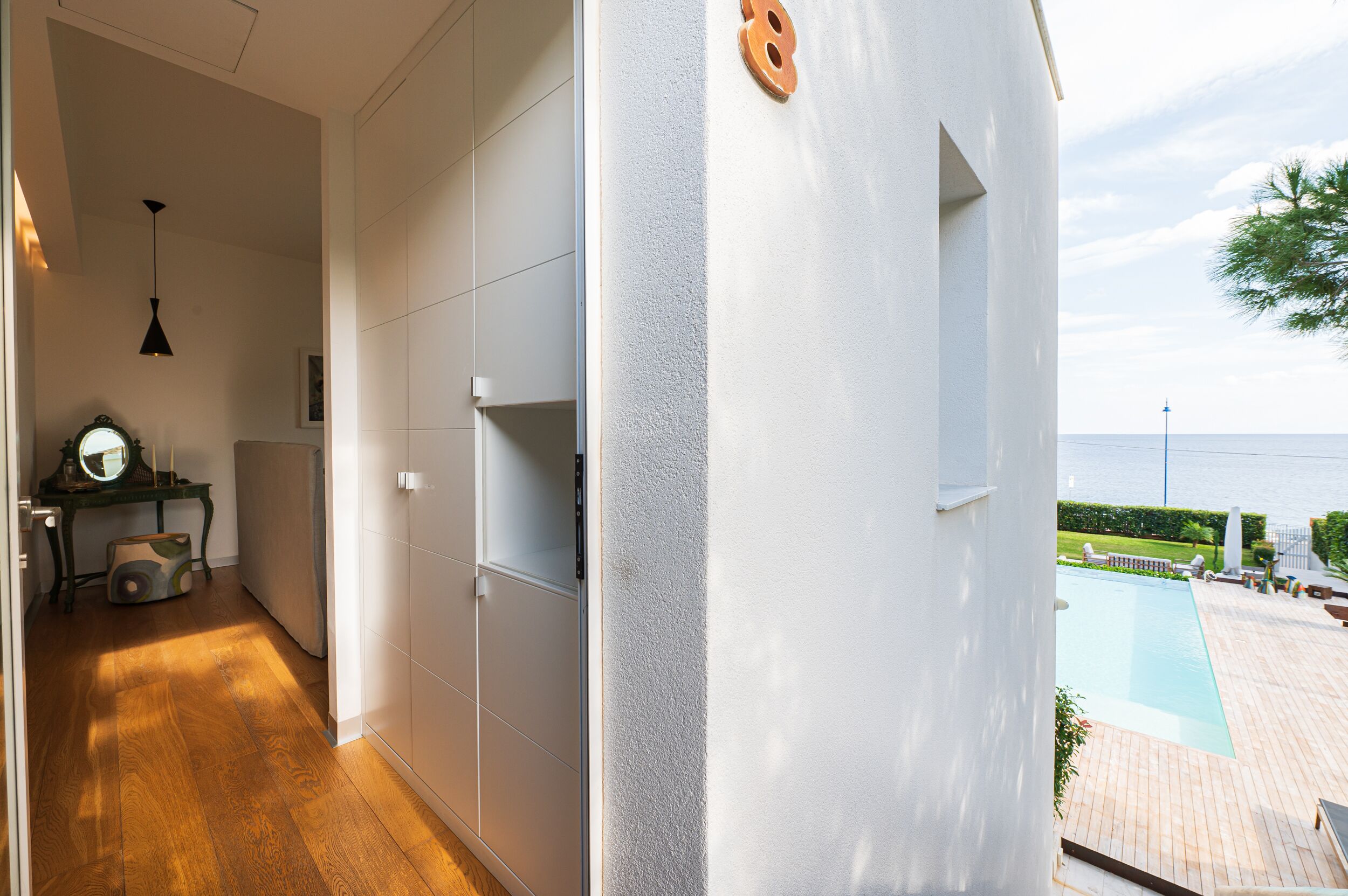 A modern interior with a wooden floor, white walls, and a view of a pool and ocean.