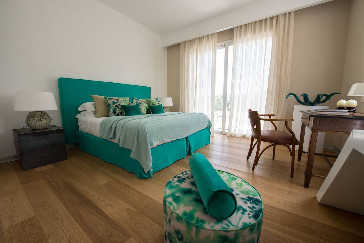 A bedroom with a teal bed, a wooden desk, a chair, and a round ottoman.