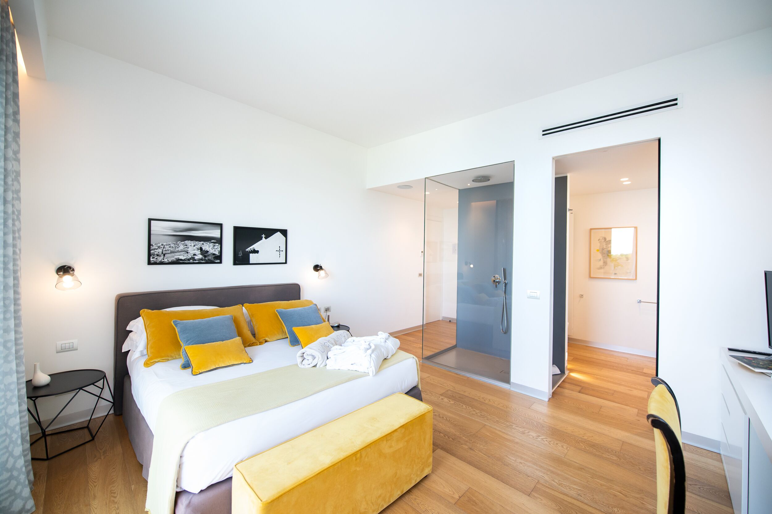 A modern hotel room with a large bed, a bedside table, a chair, a desk, and a glass-enclosed shower area.