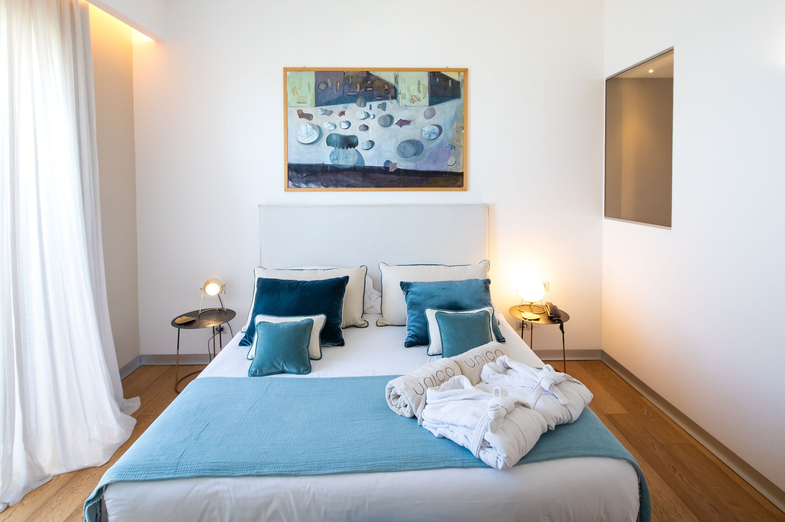 Ein ordentlich bezogenes Bett mit blauer Decke und Kissen, ein Gemälde darüber und Nachttischlampen.