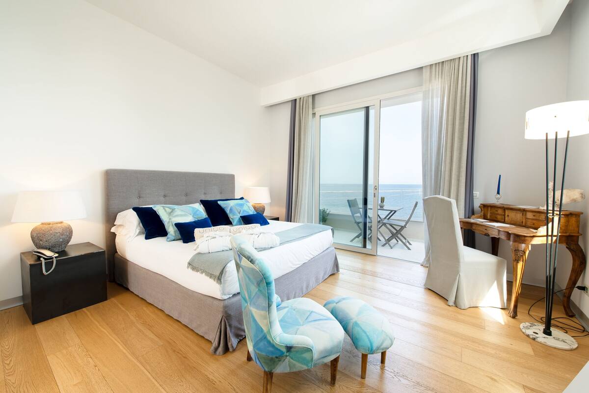A modern hotel room with a large bed, a desk, a chair, and a view of the sea.
