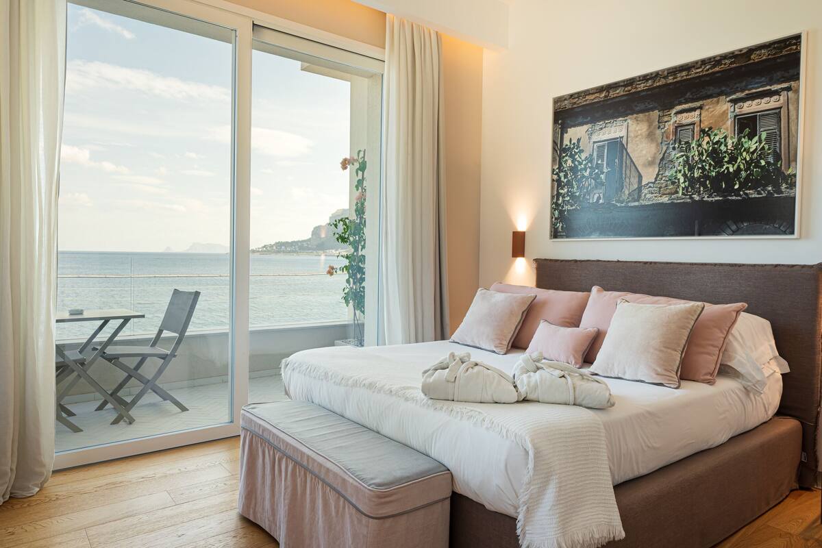 A bedroom with a large bed, a bench, a table, and chairs by a sliding glass door leading to a balcony with a view of the sea.