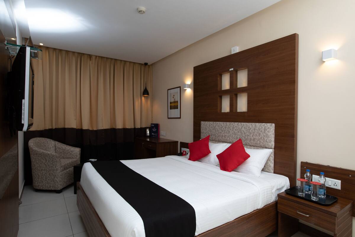 A hotel room with a large bed, a bedside table with a tray of bottled water, a chair, and a wall-mounted TV.