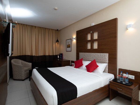 Quarto de hotel com uma cama grande, mesa de cabeceira com bandeja de água engarrafada, uma cadeira e televisão presa na parede.