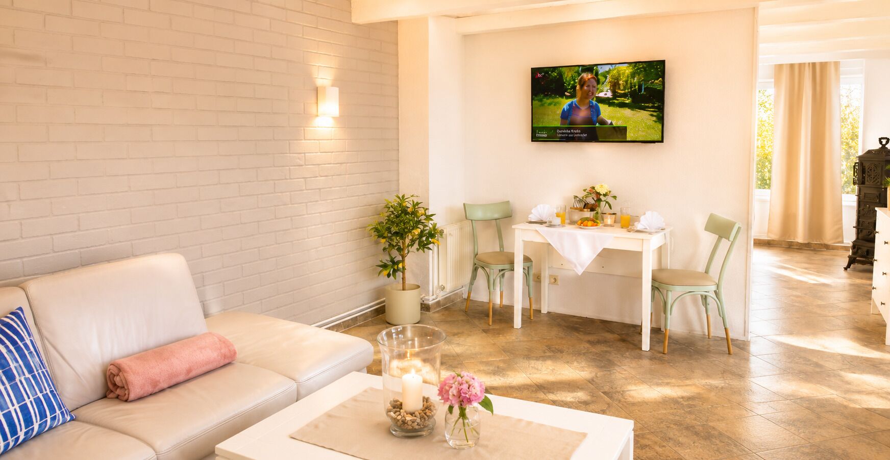 Ein Wohnzimmer mit Sofa, Couchtisch mit Kerzen und Blumen, Essbereich mit Stühlen und Tisch sowie einem an der Wand befestigten Fernseher.