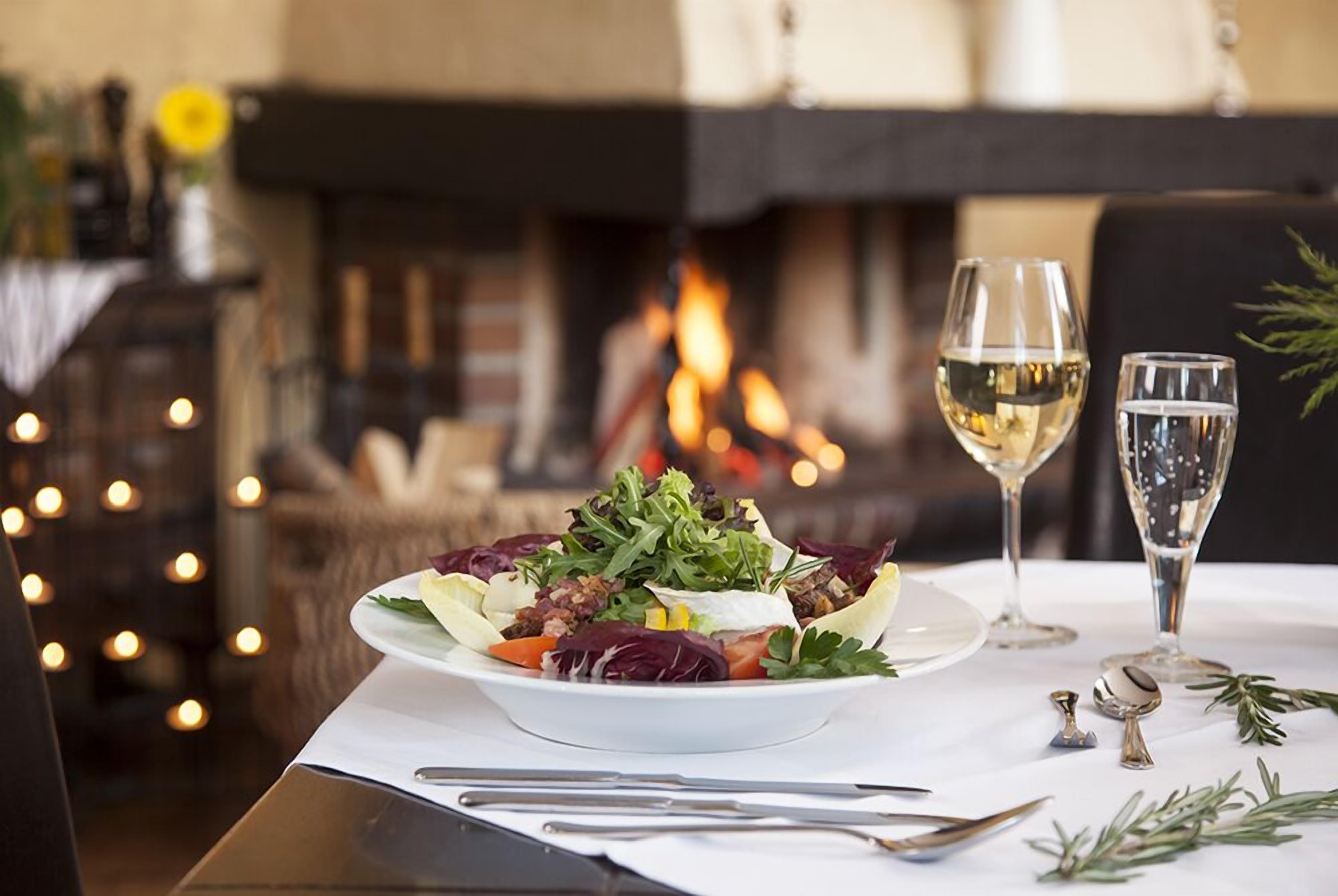Una mesa con ensalada, dos copas de vino blanco y una chimenea al fondo.