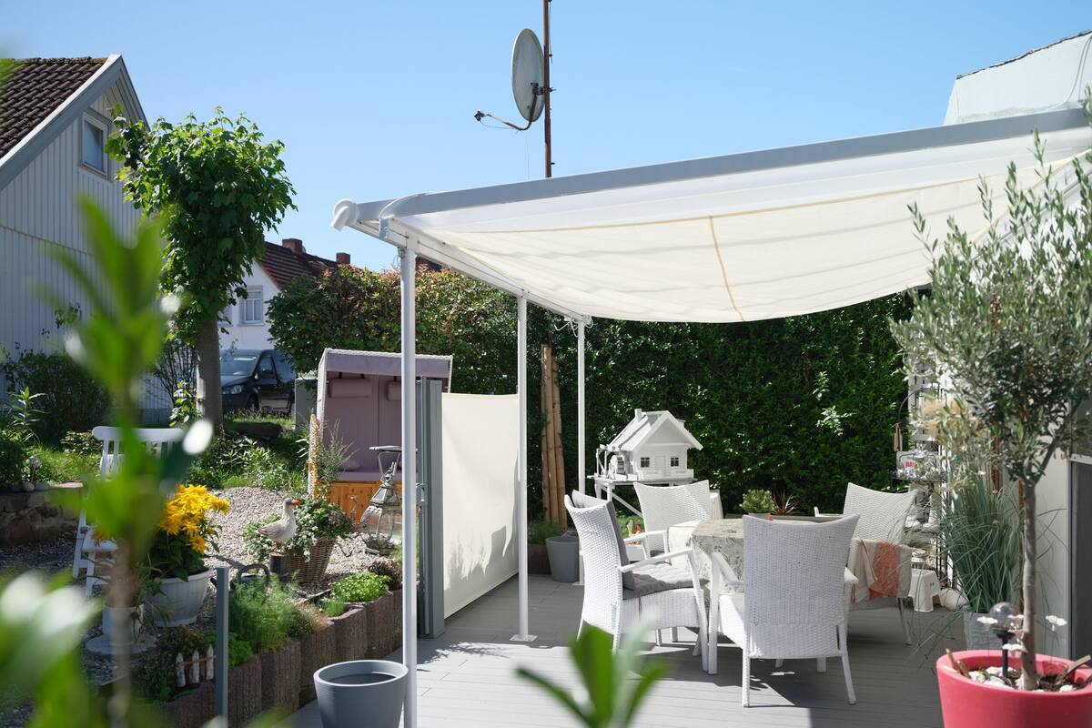 En täckt uteplats med utemöbler, en modell av ett vitt hus och krukväxter.