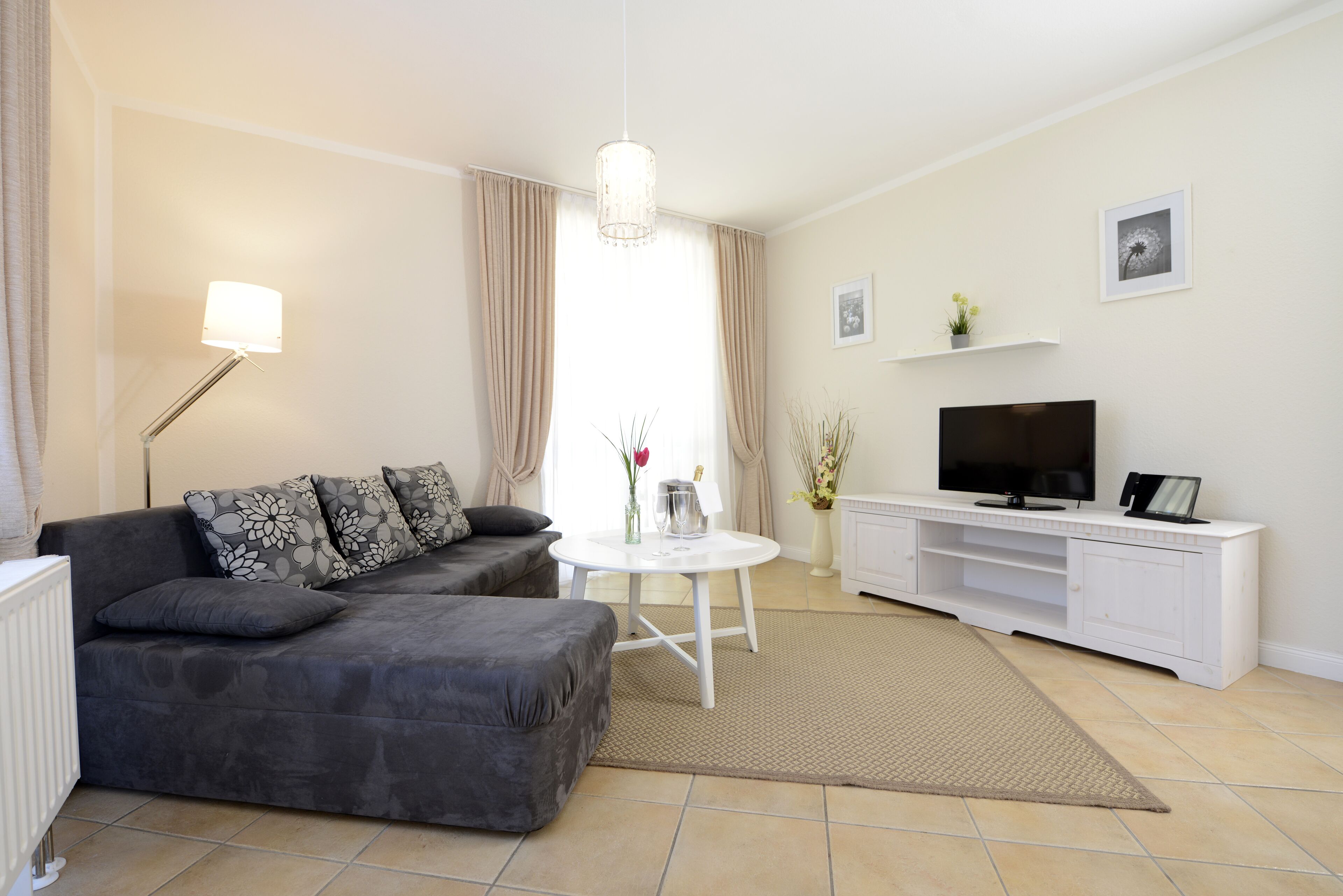 Een woonkamer met een grijze bank, een witte salontafel, een televisie op een witte kast en een radiator.