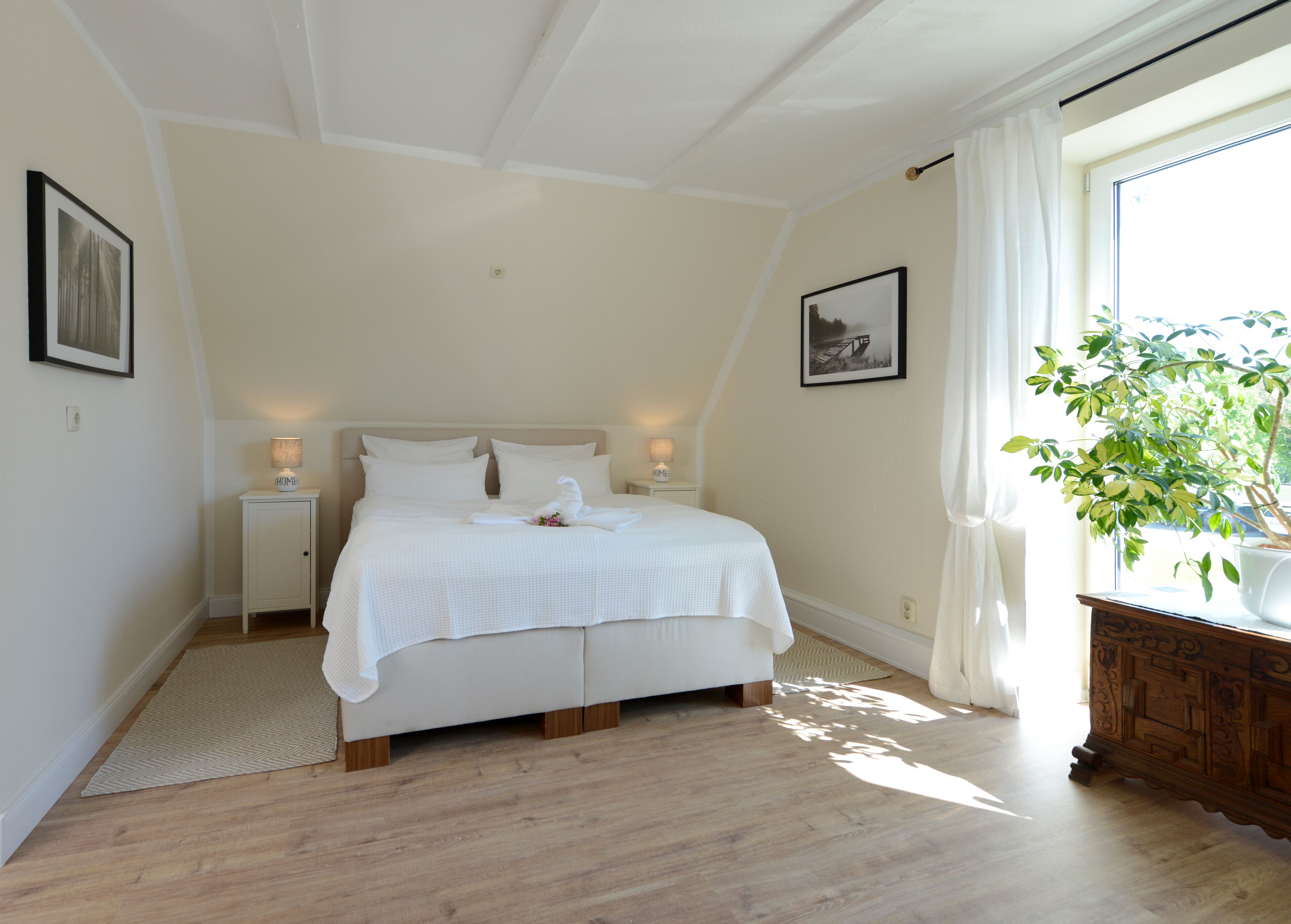 A neatly arranged bedroom with a bed, bedside tables, a nightstand, a potted plant, and a window with curtains.