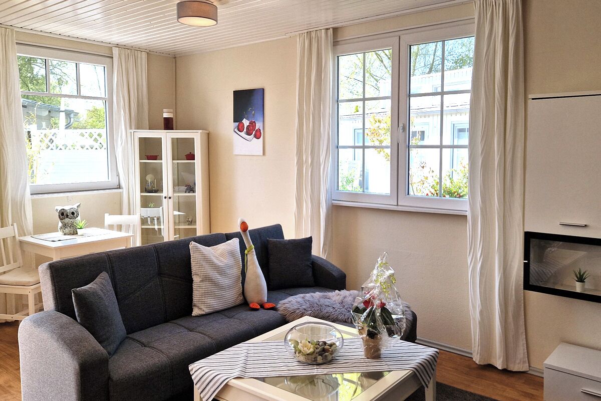 A living room with a dark grey sofa, a glass coffee table, and a white cabinet. There is a window with white curtains, a painting on the wall, and a small table with decorative items.