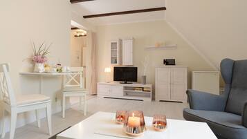 Ein modernes Wohnzimmer mit einem weißen Sofa, einem grauen Sessel, einem weißen Couchtisch mit Kerzen, einem Fernseher und einer weißen Kommode.