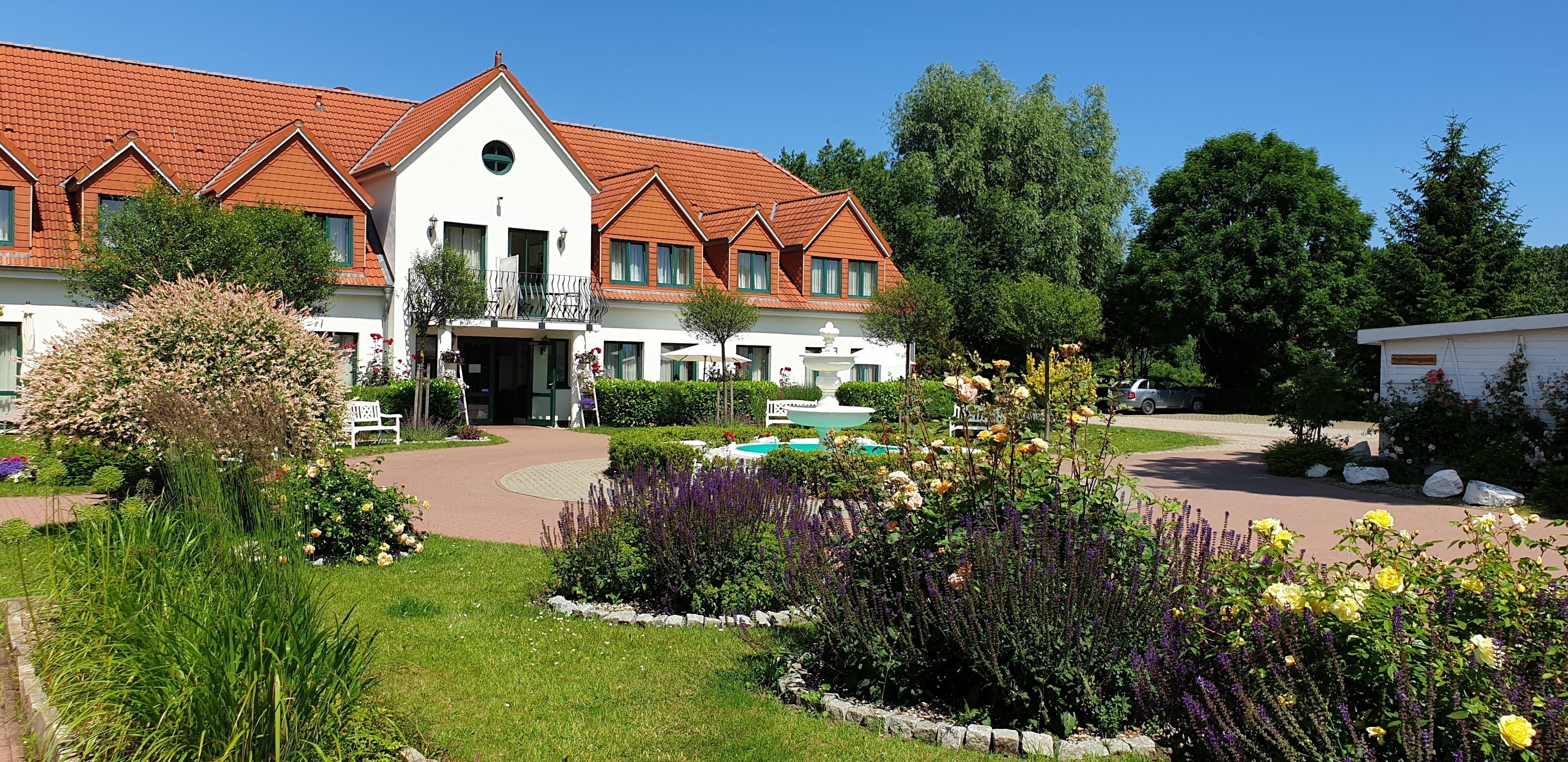 A white building with a red-tiled roof, surrounded by a well-maintained garden with various plants and flowers.