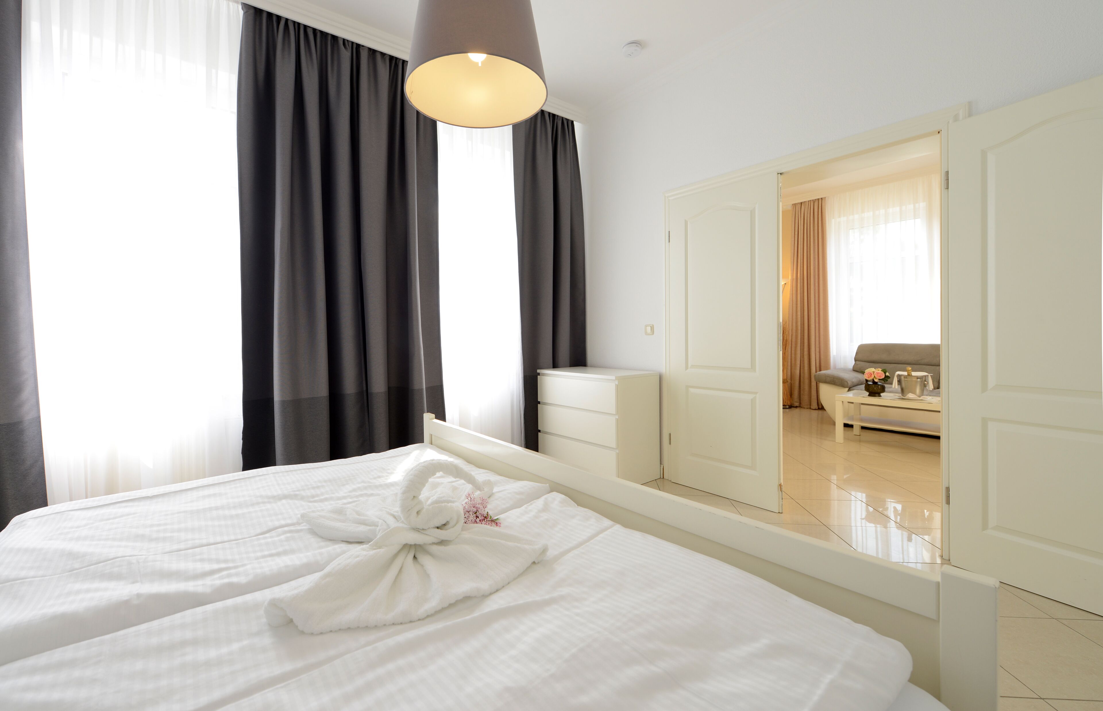 A hotel room with a large bed, a bedside table, a mirror, and a view of another room with a sofa and curtains.