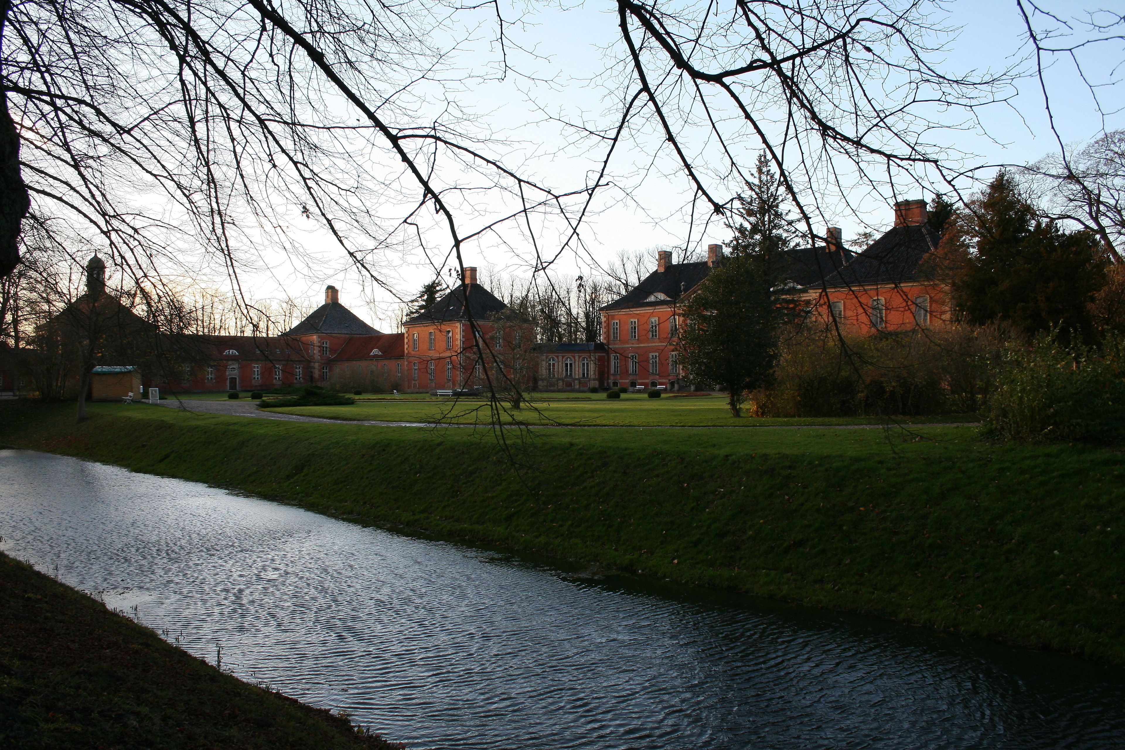 A riverside view of a historic building complex with multiple red brick structures, a grassy lawn, and a calm river.