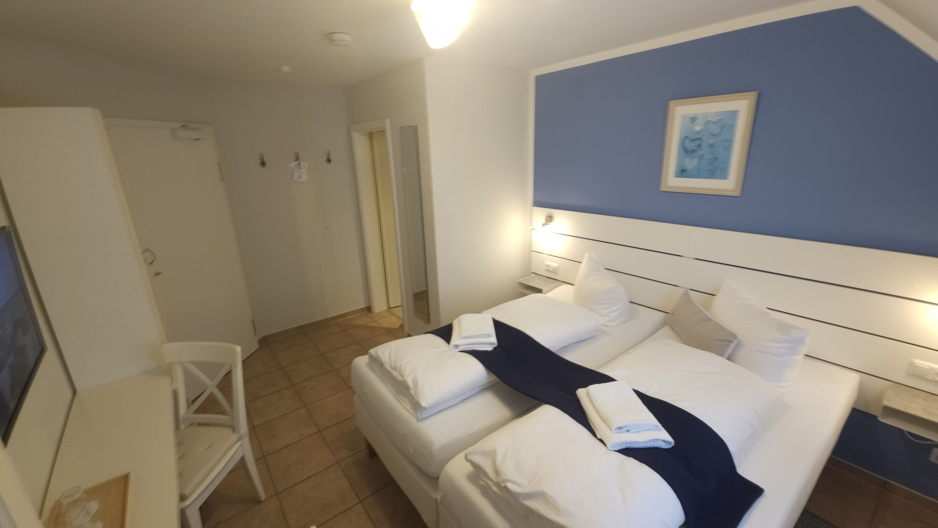 A hotel room with two beds, a desk, and a chair. There is a painting on the wall and a door leading to another room.