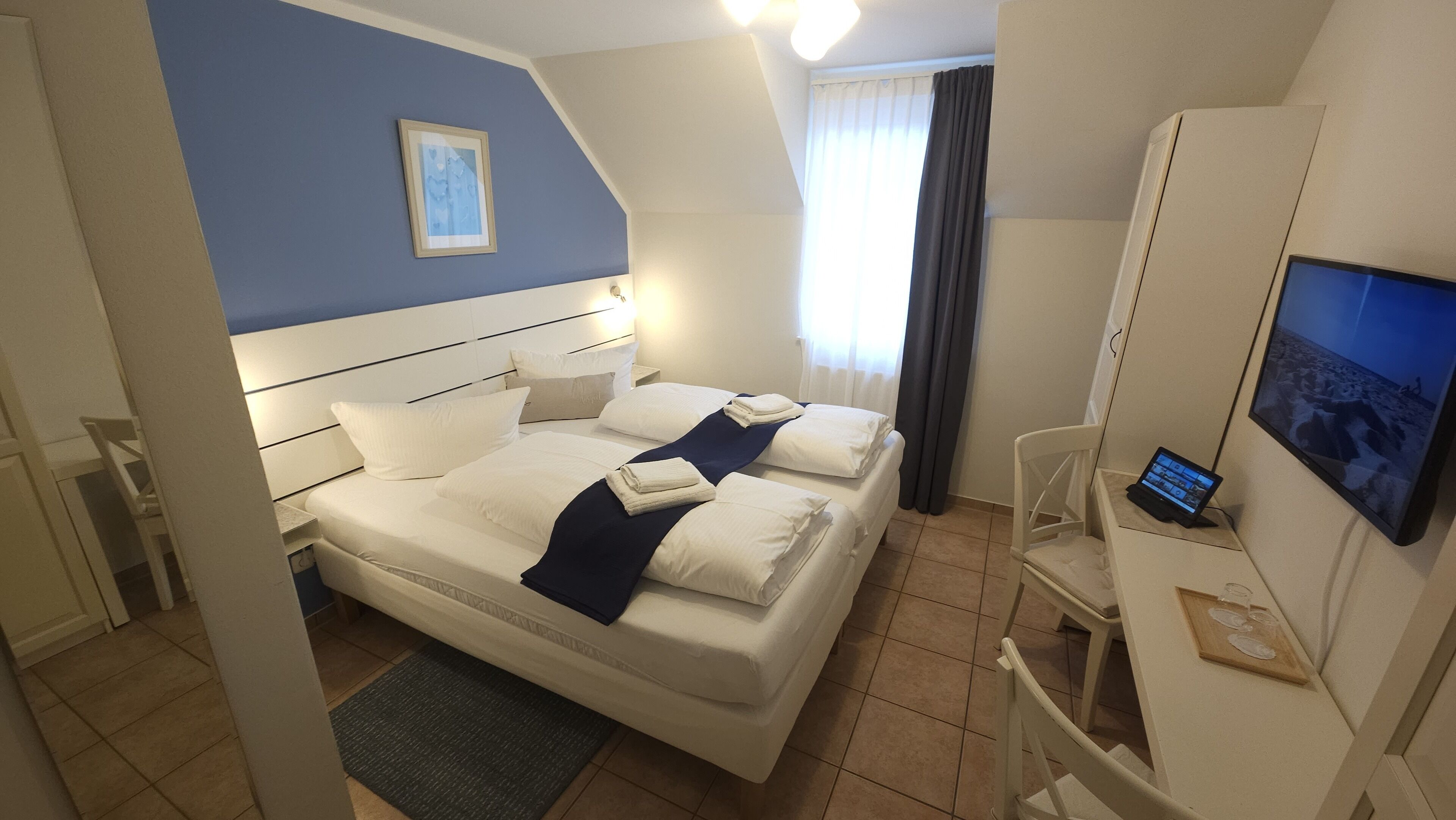 Quarto de hotel com uma cama, uma escrivaninha com um laptop, uma televisão e uma janela com cortinas.