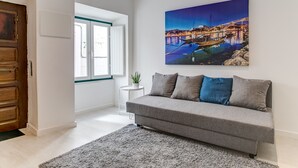 A modern living room with a grey sofa, a small white side table with a plant, and a large painting of a bridge over water.
