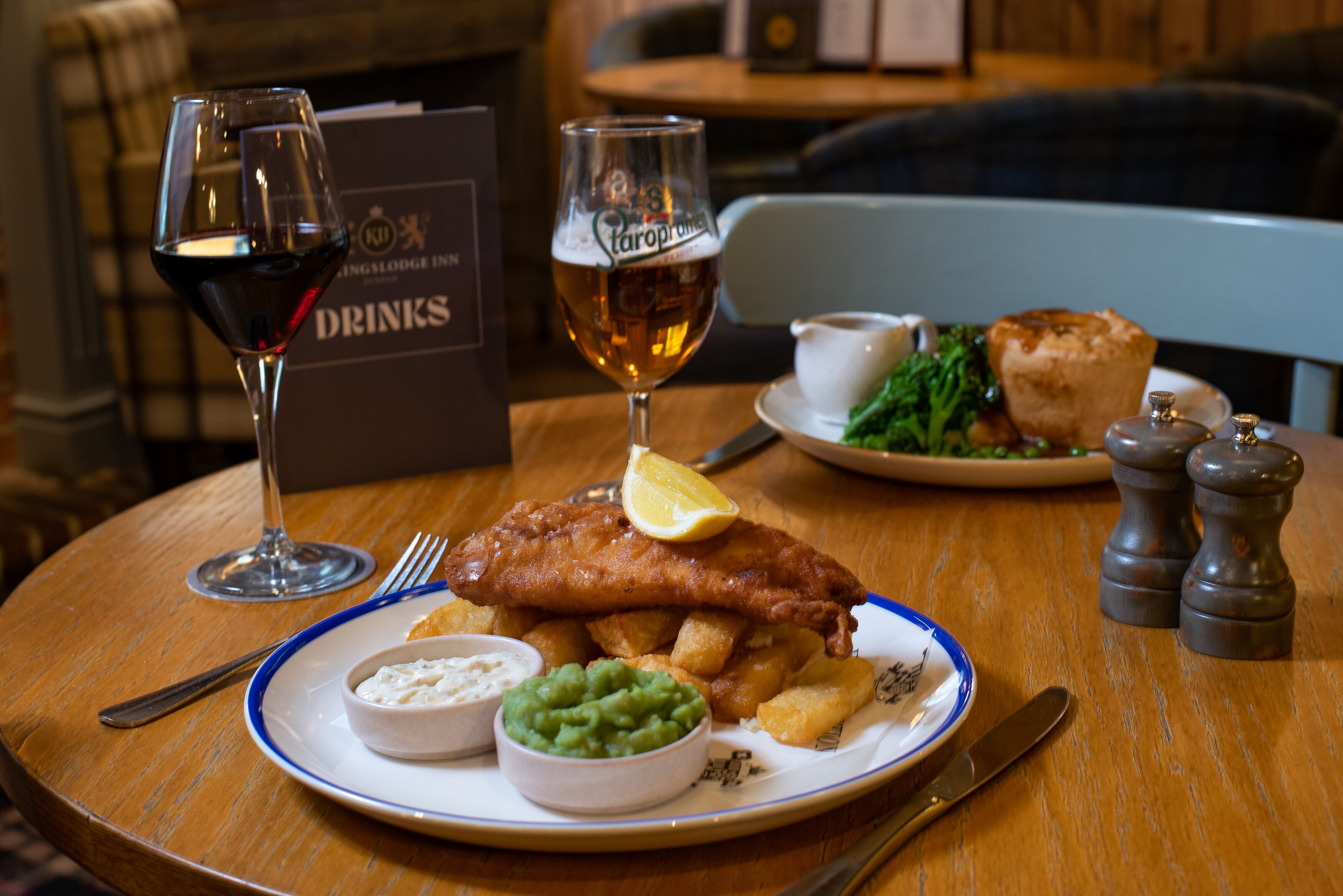Une assiette de poisson-frites, accompagnée d’un verre de bière et d’un verre de vin rouge, sur une table en bois.