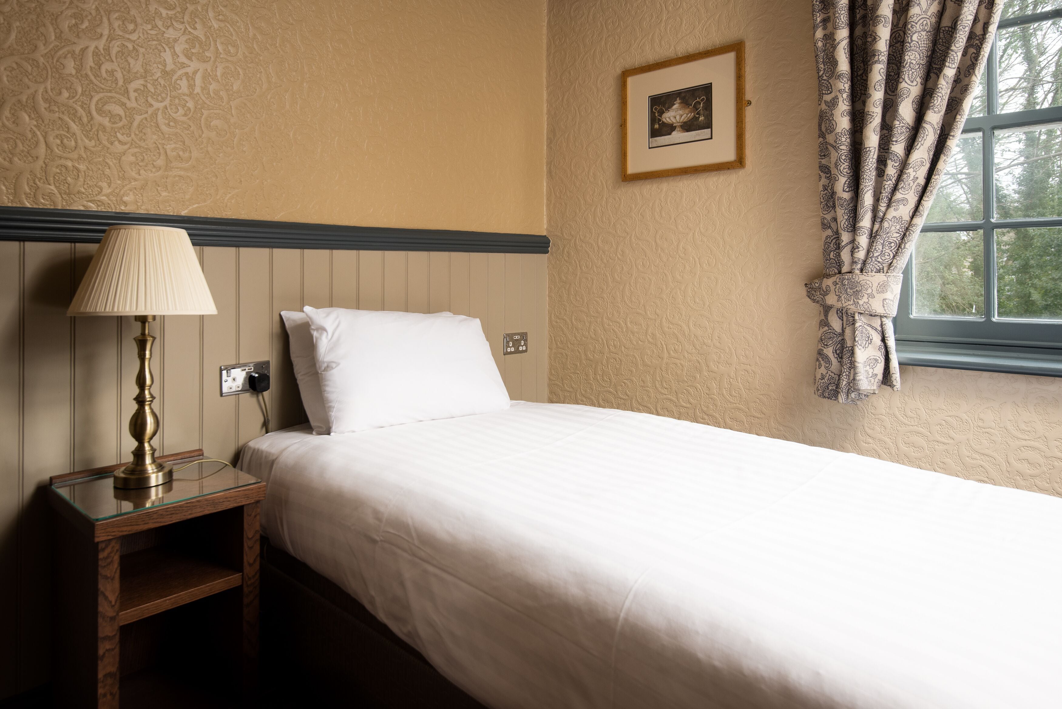 A neatly made bed with a white comforter and pillows, a bedside table with a lamp, and a window with patterned curtains.