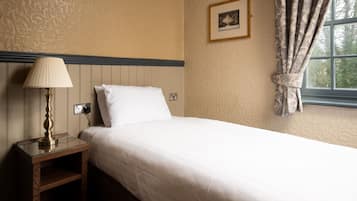 A neatly made bed with a white comforter and pillows, a bedside table with a lamp, and a window with patterned curtains.