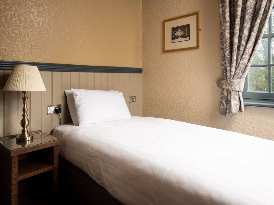 A neatly made bed with a white comforter and pillows, a bedside table with a lamp, and a window with patterned curtains.