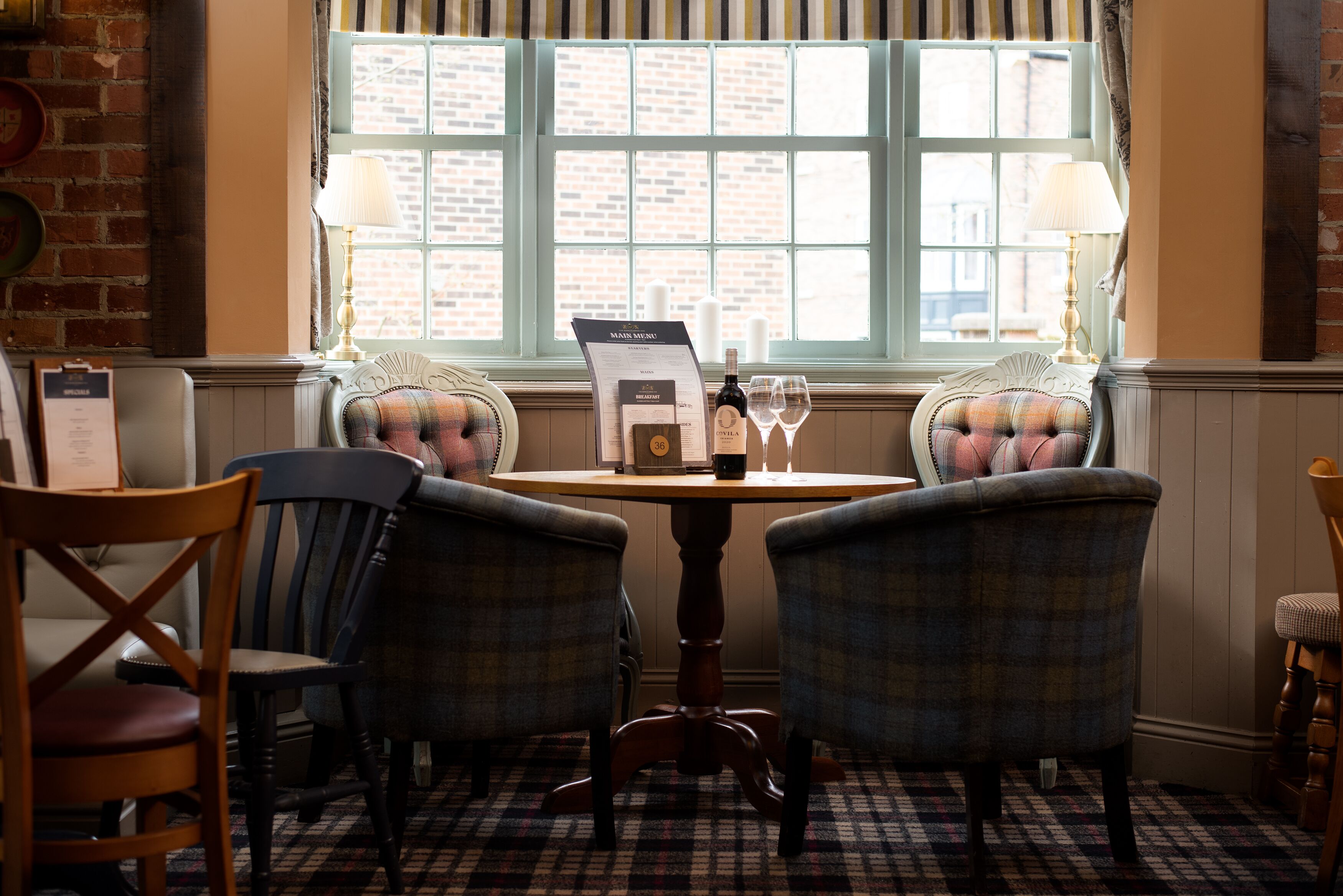A dining area with a table, two plaid chairs, a menu stand, and a window with a view of a brick building.