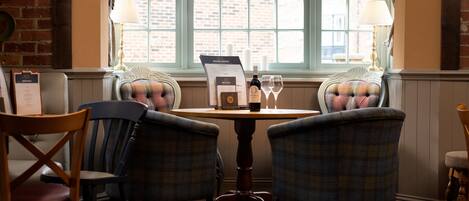 A dining area with a table, two plaid chairs, a menu stand, and a window with a view of a brick building.