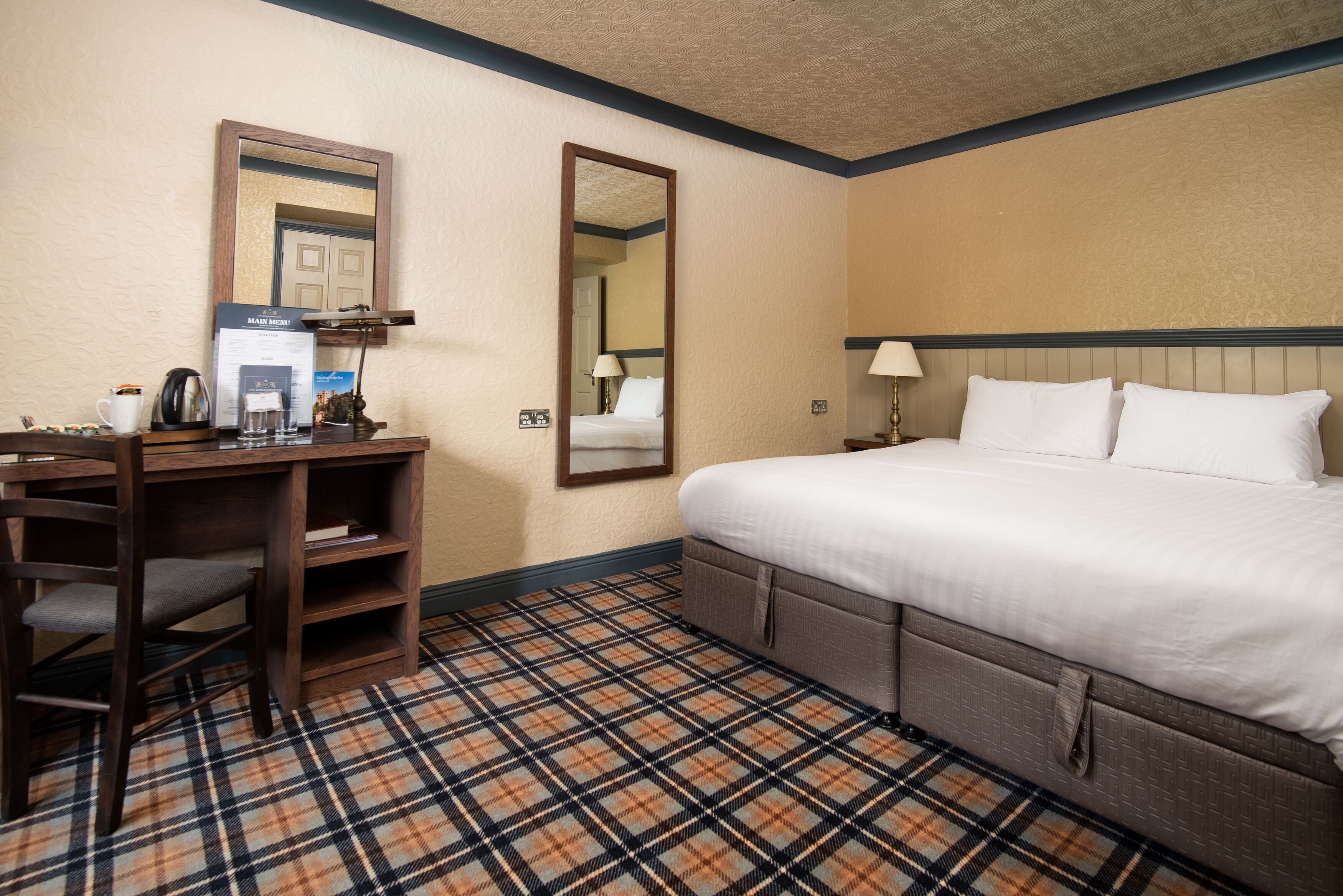 A hotel room with a bed, a desk with a chair, a mirror, and a check-in counter.
