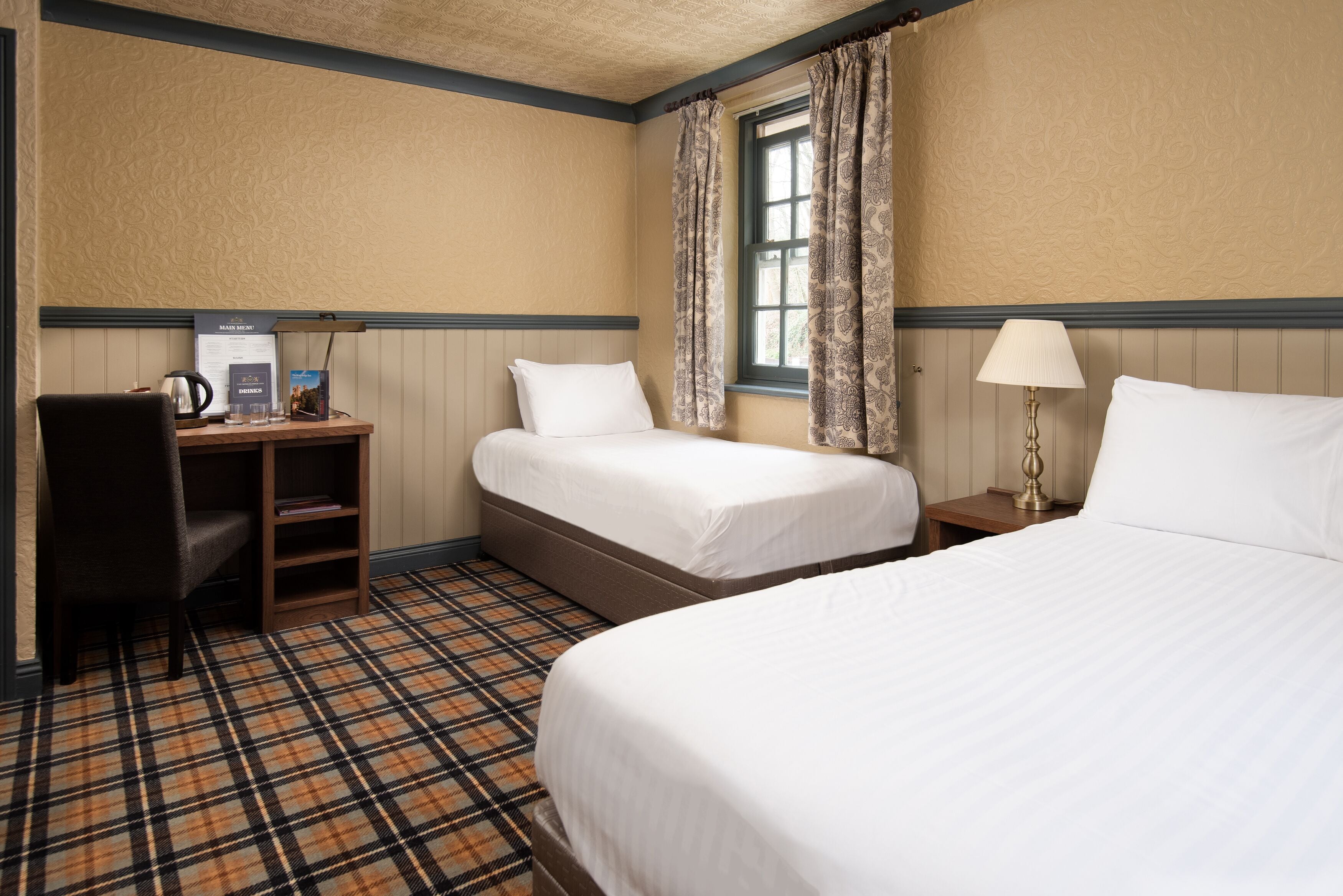 A hotel room with two beds, a desk with a chair, a lamp, and a window with curtains.