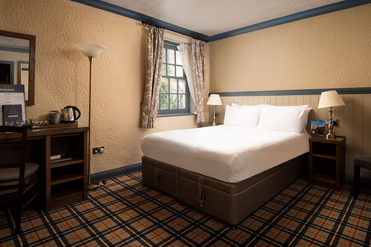 A hotel room with a large bed, a desk with a lamp, a window with curtains, and a plaid-patterned floor.
