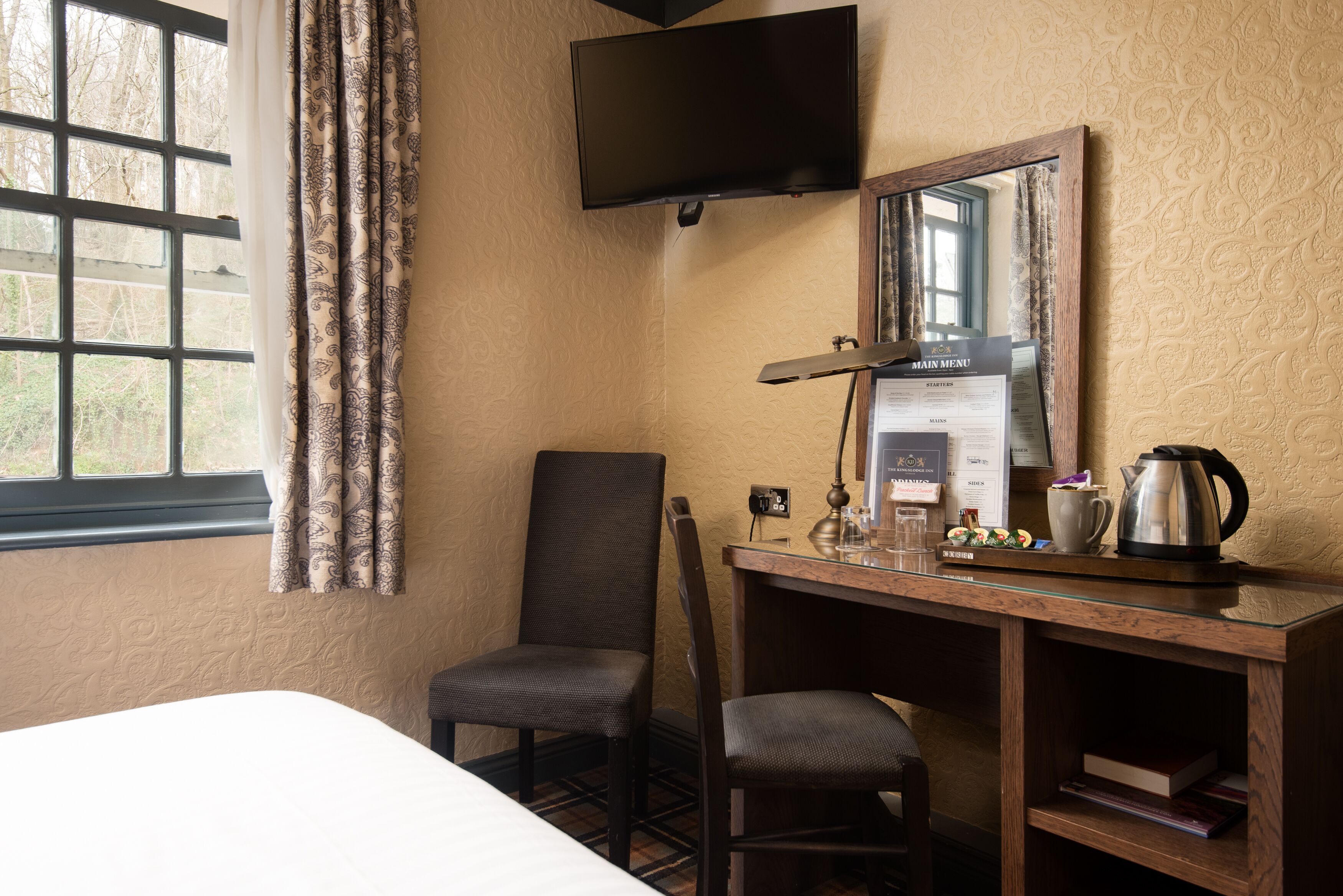 A hotel room with a bed, a desk with a computer, a chair, a TV mounted on the wall, and a window with curtains.