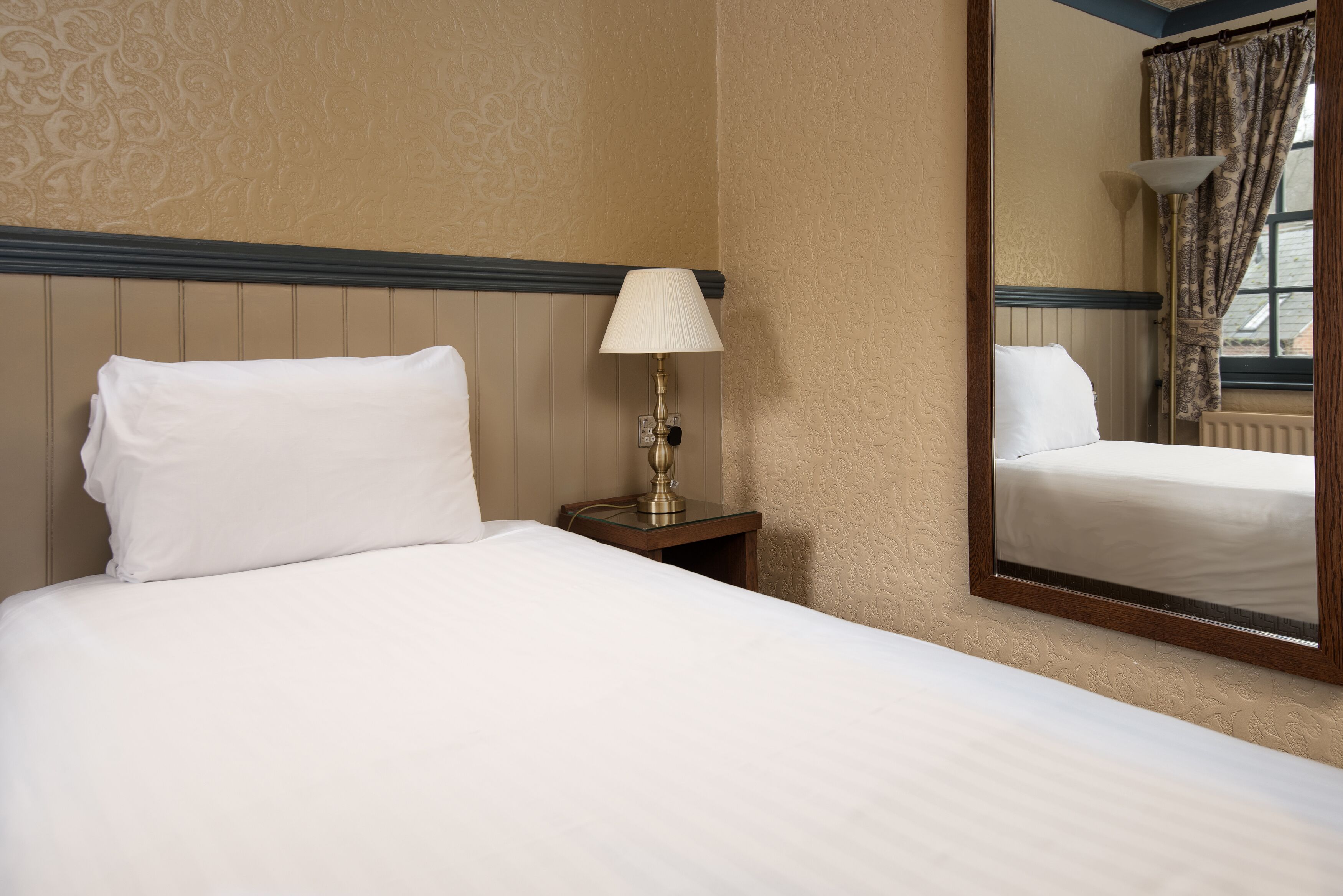 Una cama bien hecha con sábanas blancas, una lámpara de noche y un espejo que refleja la habitación.